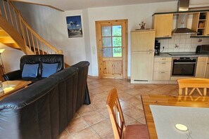 A living room with a black leather sofa, a wooden dining table with chairs, a kitchen area with cabinets and appliances, and a wooden staircase. - Holiday home with 3 bedrooms (Ostseebad Prerow)