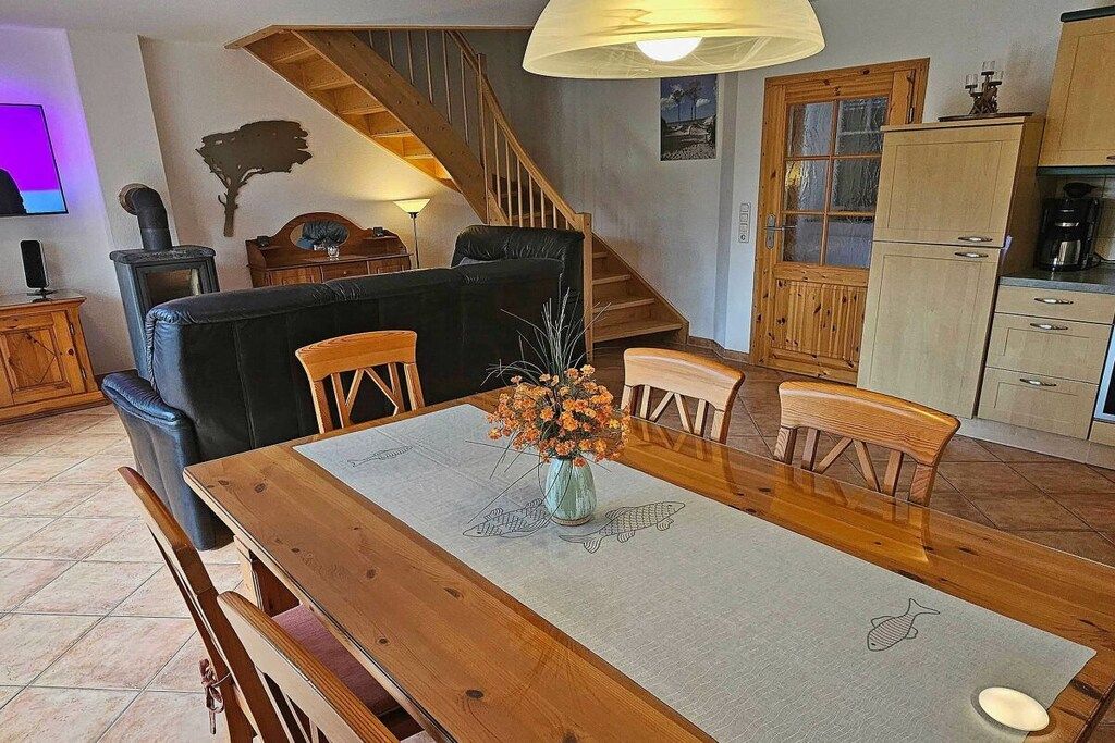 Un coin repas avec une table en bois, des chaises et un vase de fleurs. On aperçoit un escalier au fond.