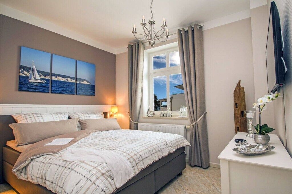 A bedroom with a bed, a bedside table with a lamp, a window with curtains, and a wall-mounted TV.