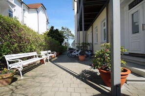 A paved walkway with benches and potted plants, leading to a building with white doors and windows. - Beach oasis - 1st row by the sea (Ostseebad Binz)