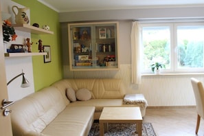 A living room with a beige sofa, a small table, a bookshelf, and a window with a view of greenery. - Holiday apartment with 1 bedroom (Dresden)