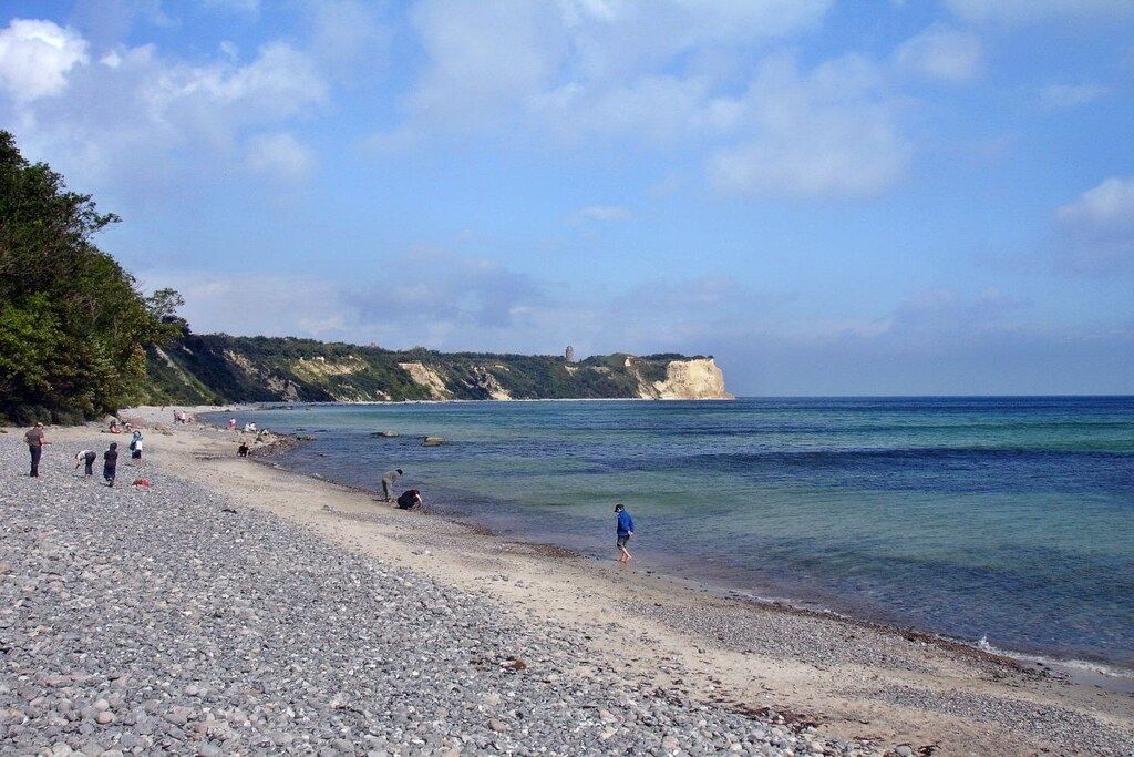 Una playa de guijarros con gente caminando y parada, un cielo despejado y un acantilado distante.