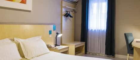 A hotel room with a bed, bedside table, lamp, air conditioning unit, and a desk with a chair.