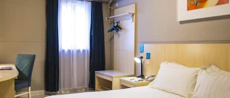 A hotel room with a bed, a desk, a chair, an air conditioning unit, a painting, and a window with curtains.