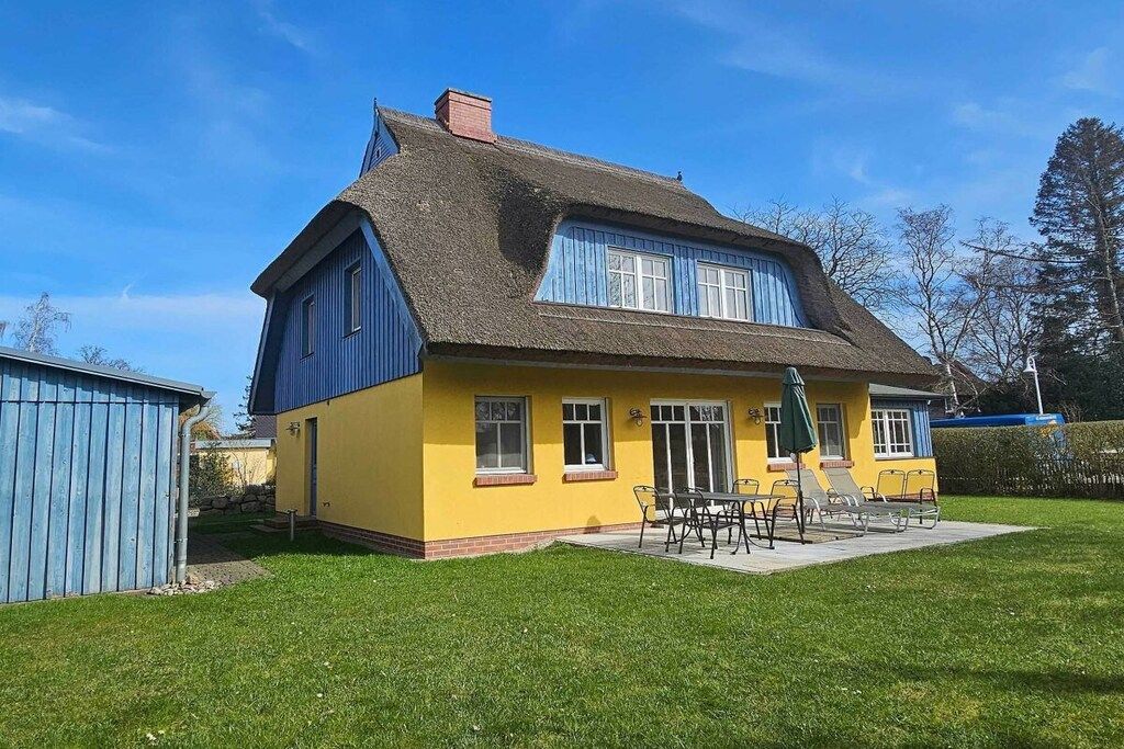 A two-story house with a thatched roof, a yellow facade, and a patio with outdoor furniture.