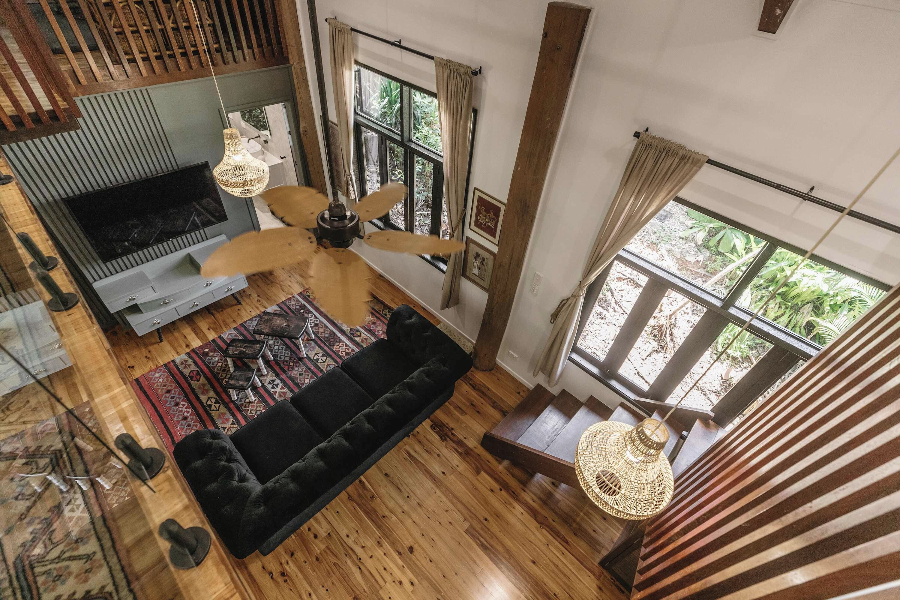 Ein großzügiger Wohnbereich mit schwarzem Sofa, Holzboden, Deckenventilator und großen Fenstern mit Blick auf Grünflächen.