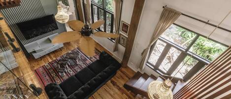 Ein großzügiger Wohnbereich mit schwarzem Sofa, Holzboden, Deckenventilator und großen Fenstern mit Blick auf Grünflächen.