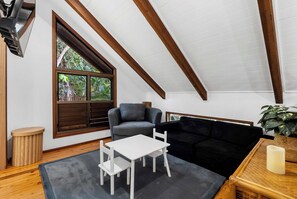 A cozy room with a sloped ceiling, a window with wooden shutters, a grey sofa, a white table, and a black sofa. - Treehouse—Serene Tropical Hideaway & Private Pool (Smithfield)
