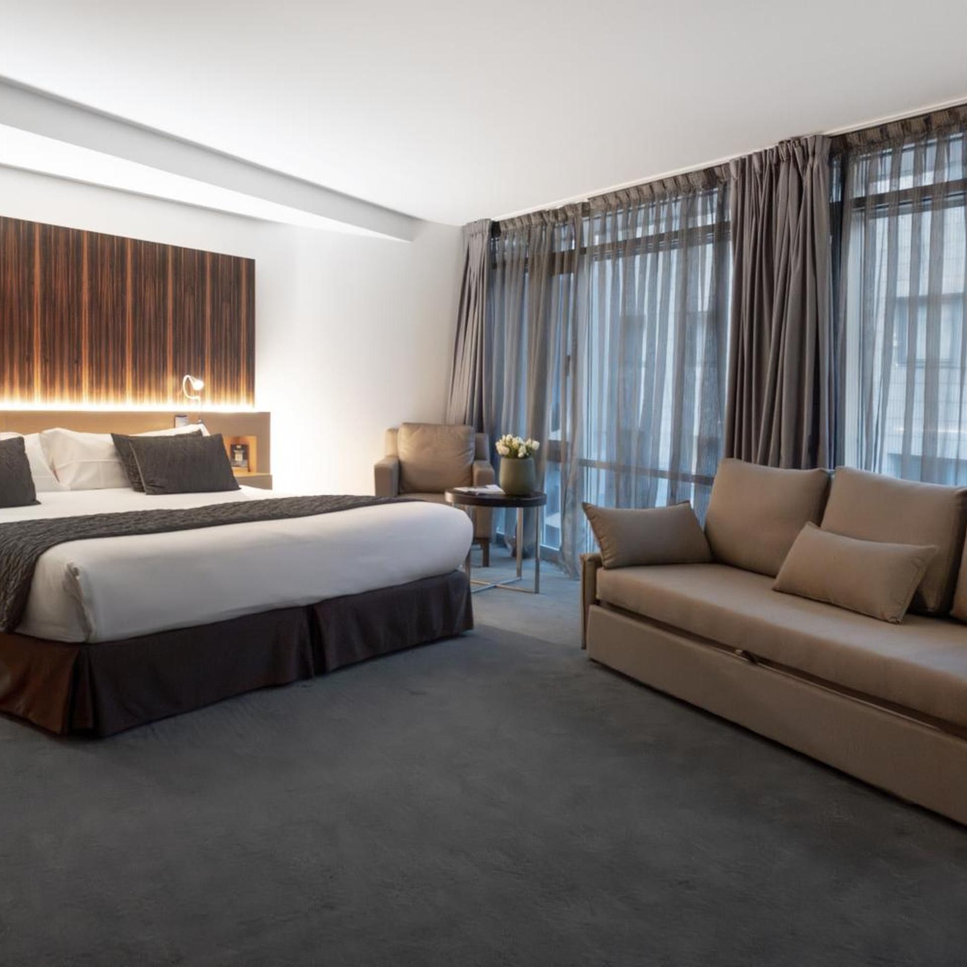 A modern hotel room with a large bed, a sofa, a small table with a vase, and sheer curtains.