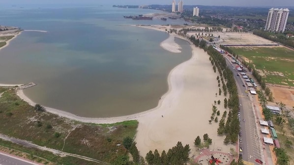 An aerial view of a coastal area with a sandy beach, a road lined with trees, and buildings in the background.