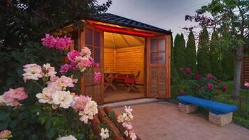 A wooden cabin with a bench and a table outside, surrounded by a garden with roses.