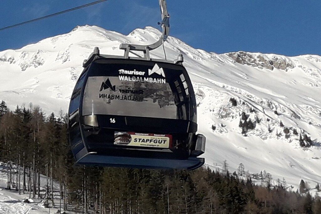A chairlift with 'Rauriser Waldalmbahn' and 'Stapfgut' logos, ascending a snowy mountain.