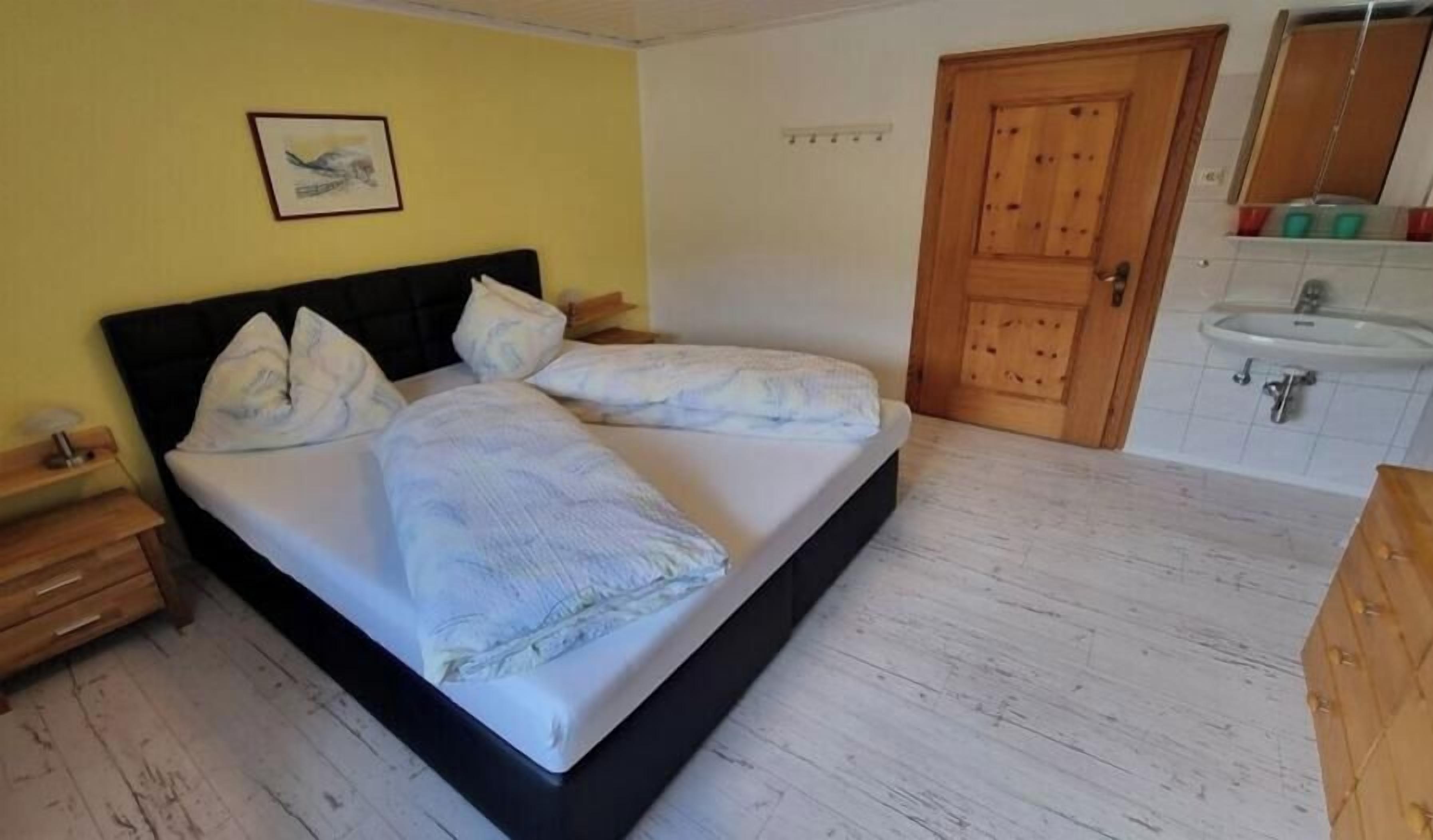 A bedroom with a bed, bedside table, wooden wardrobe, and a bathroom with a sink and mirror.