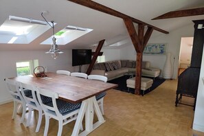 Un coin repas avec une table en bois et des chaises blanches, un téléviseur et un canapé à l’arrière-plan.