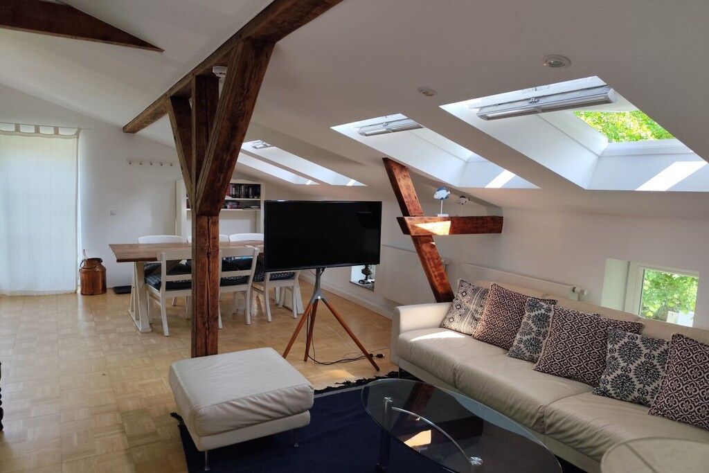 A spacious loft with a high ceiling, skylights, a sofa with patterned cushions, a flat-screen TV, and a dining area with a table and chairs.