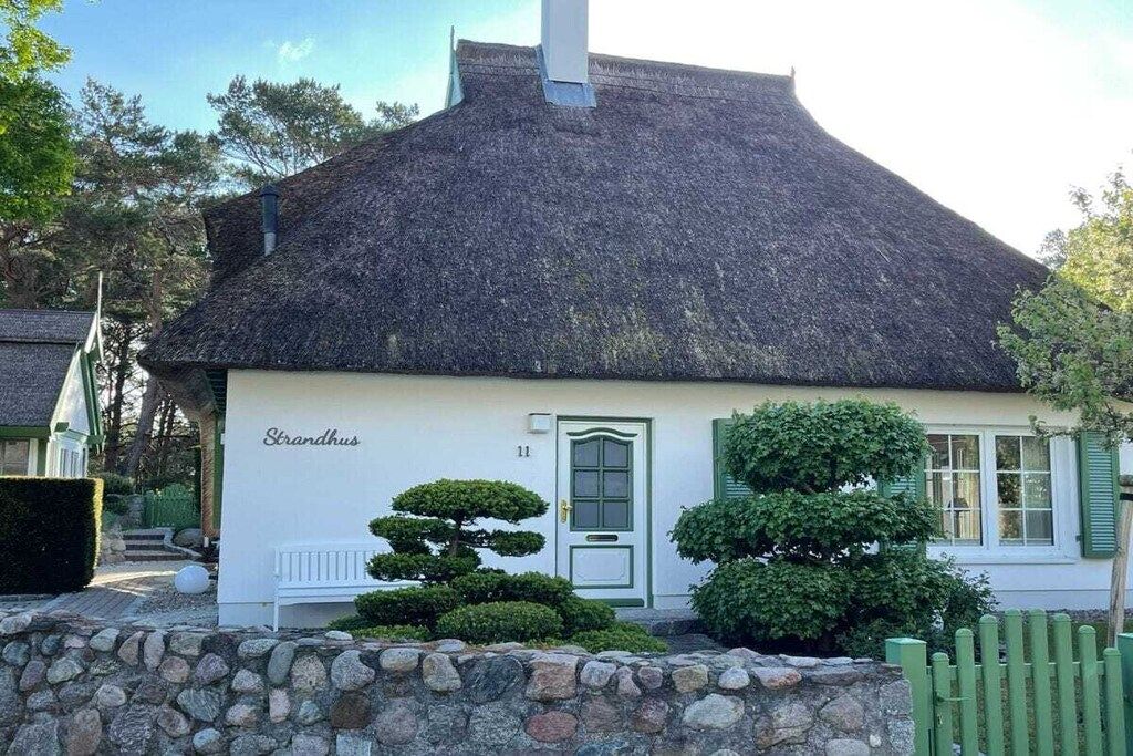 Uma casa tradicional com telhado de palha, um muro de pedra em frente e um arbusto bem aparado.
