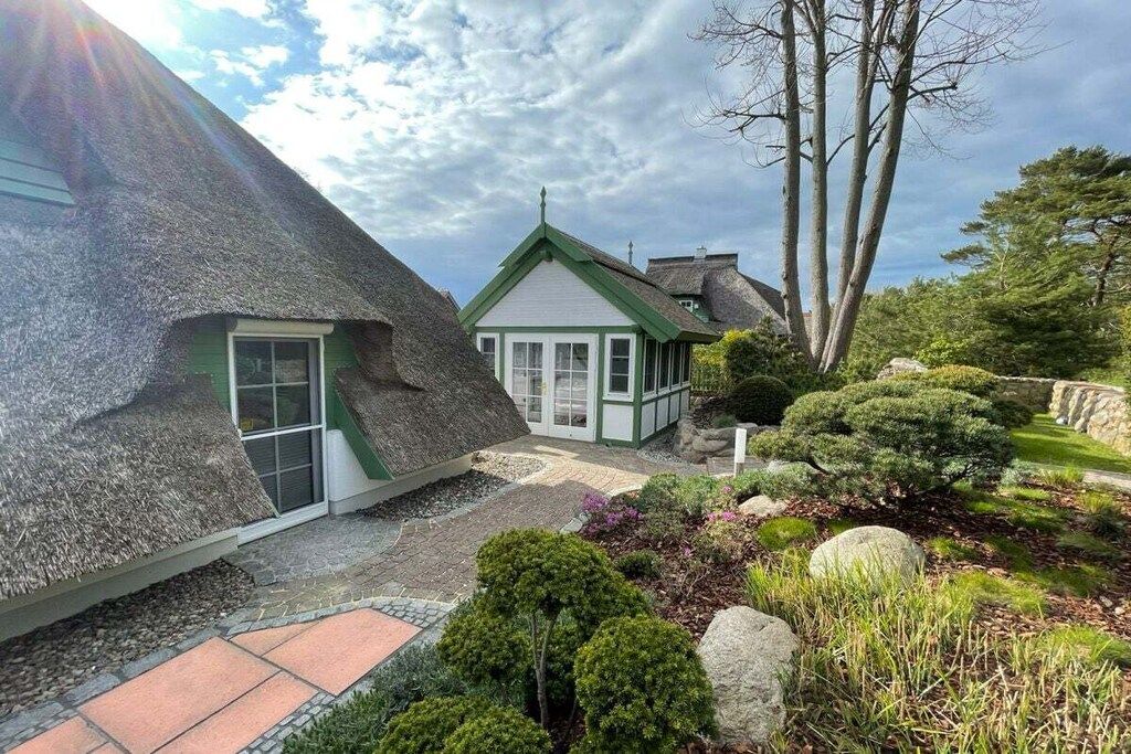Een traditioneel rieten huis met een klein aanbouwtje, omgeven door een verzorgde tuin met verschillende struiken en een pad van stenen.