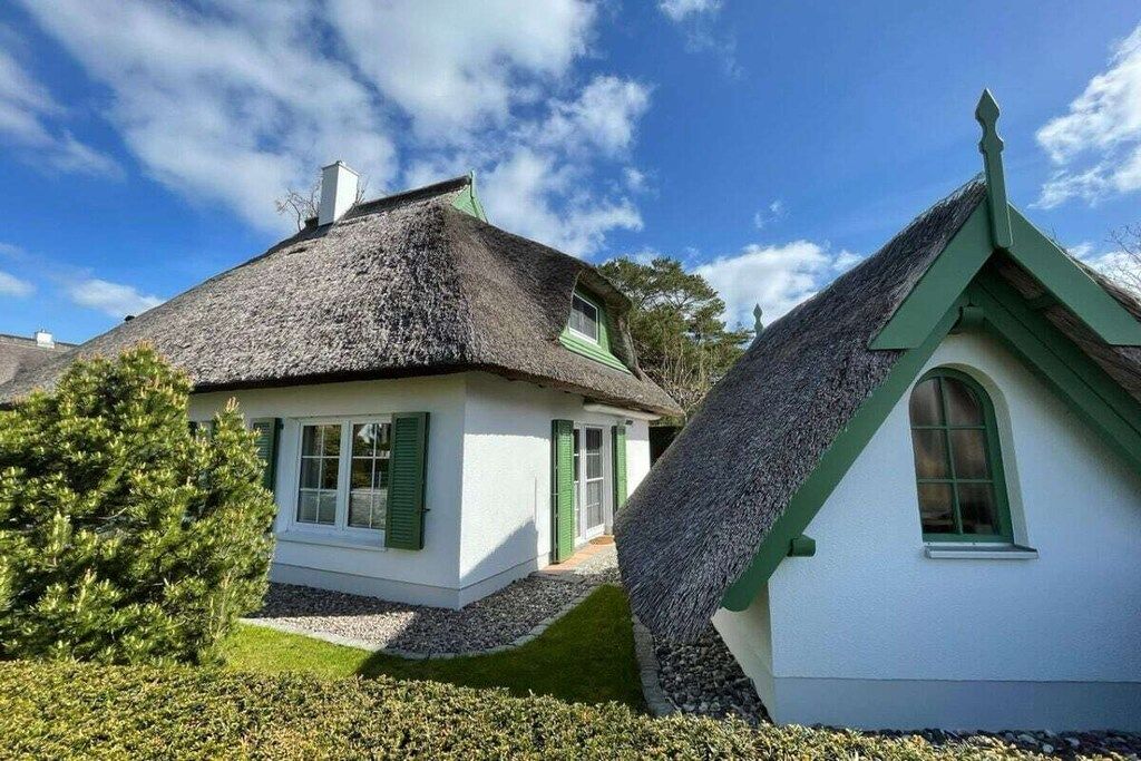 Una casa tradicional con techo de paja y fachada blanca, con contraventanas verdes, rodeada de setos cuidados y un cielo despejado.
