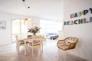 Ein helles Wohnzimmer mit Essbereich, einem Korbstuhl und einer Wand mit der Aufschrift 'HAPPY BEACHL!'.