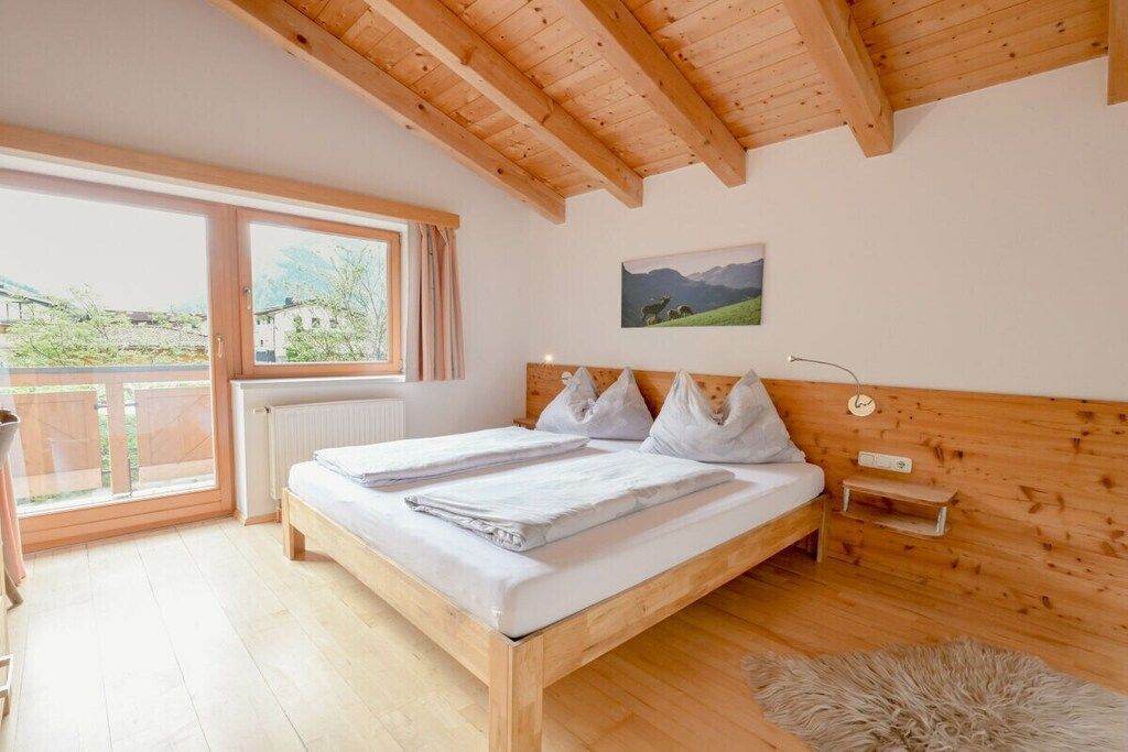A bedroom with a wooden ceiling, a large window, a bed with white linens, a small bedside table, and a wall-mounted lamp.