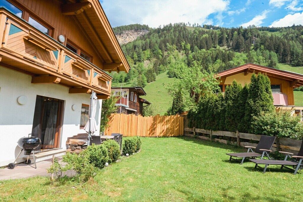 A wooden chalet with a balcony, a barbecue, and a fenced backyard with lounge chairs.
