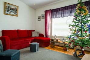 A living room with a red sofa, a small table, a Christmas tree, and a window with curtains.