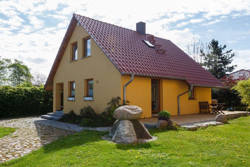 A two-story house with a red-tiled roof, a stone pathway leading to the entrance, and a grassy yard with a large rock feature.