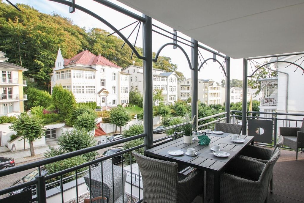 Un balcón con mobiliario de mimbre y una mesa preparada para comer, con vistas a una zona residencial con edificios y árboles.
