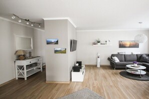 Una sala de estar moderna con sofá, mesa de centro y televisor. Se observa una consola blanca con una lámpara y un estante con adornos.