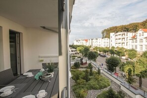 A balcony with a table set for dining, overlooking a landscaped garden and a street with buildings. - Villa Louisa 40 - Lighthouse (Ostseebad Sellin)