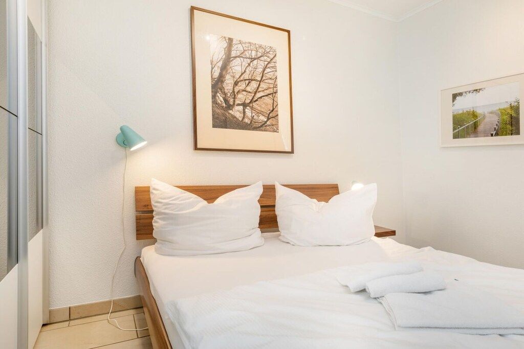 Ein ordentlich bezogenes Bett mit weißen Leinen und Kissen, ein gerahmtes Bild darüber und eine Wandlampe links.