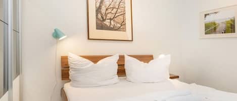 Un lit bien fait, avec du linge de lit blanc et des oreillers, une photo encadrée au-dessus, et une lampe fixée au mur sur le côté gauche.