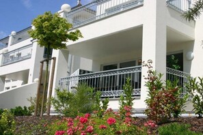 A white building with balconies, a tree, and flowering plants. - Dune Residence Queen Augusta 5 (Ostseebad Binz)