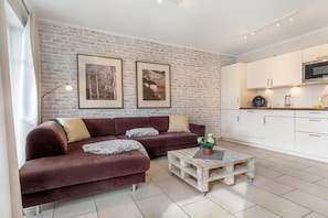 A living room with a sofa and a coffee table, a kitchen with white cabinets, and a brick wall. - Dune Residence Queen Augusta 5 (Ostseebad Binz)