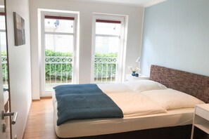 A bedroom with a bed, two windows, a nightstand, and a view of greenery outside. - Dune Residence Queen Augusta 3 (Ostseebad Binz)