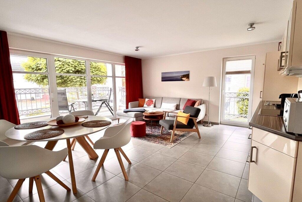 A modern living room with a sofa, coffee table, and chairs. There is a dining area with a table and chairs. The room has large windows and a view of the outdoors.