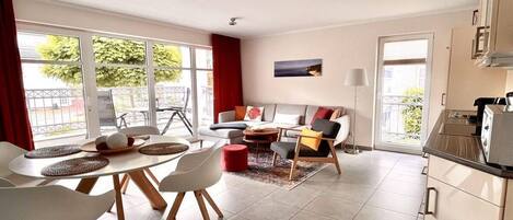A modern living room with a sofa, coffee table, and chairs. There is a dining area with a table and chairs. The room has large windows and a view of the outdoors.