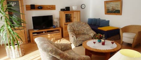 En stue med fjernsyn, to lænestole, en sofa, et lille bord med lampe og en stor plante.