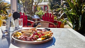 A dining table with a variety of foods, including meats, vegetables, and fruits, set outdoors with a red chair and a metal bucket in the background. - The Factory (Hapuku)
