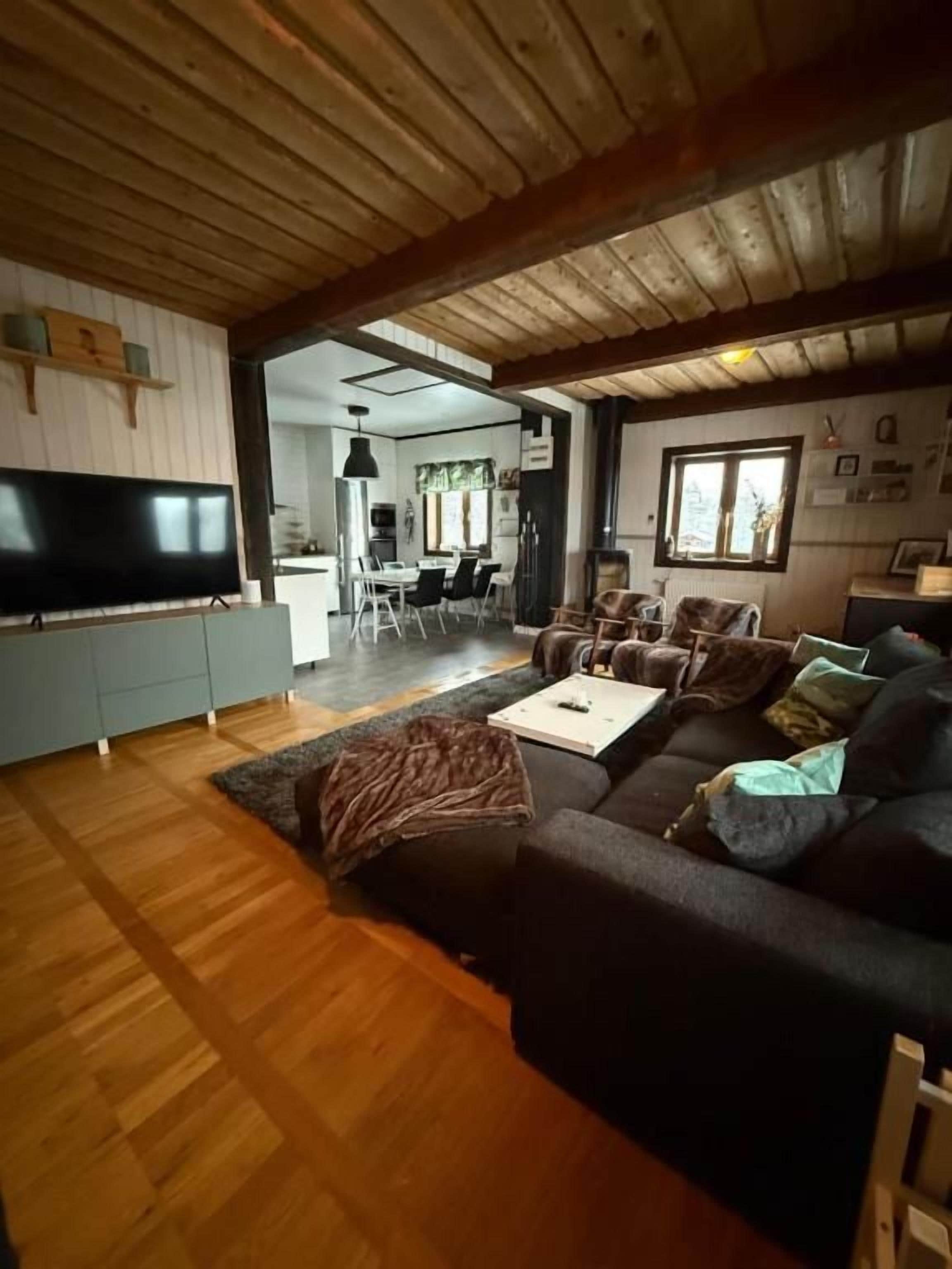 A living room with a wooden ceiling, a large flat-screen TV, a dark sofa, and a dining area in the background.
