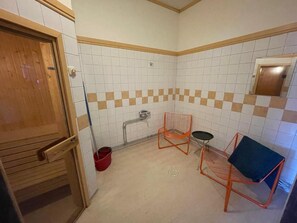 Un baño pequeño con paredes de azulejos blancos, una puerta de madera, un cubo rojo y dos sillas de metal.