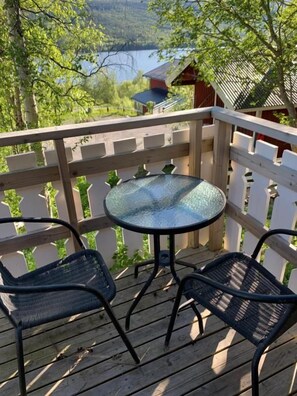Una terraza de madera con una mesa de vidrio y dos sillas, con vista a un lago y un área boscosa.