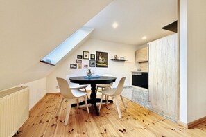 Ein Essbereich mit einem runden Tisch, weißen Stühlen und einer schwarzen Tischdecke. Über dem Tisch befindet sich ein Oberlicht, und im Hintergrund ist eine Küche mit Holzausbau und einer schwarzen Arbeitsplatte zu sehen.