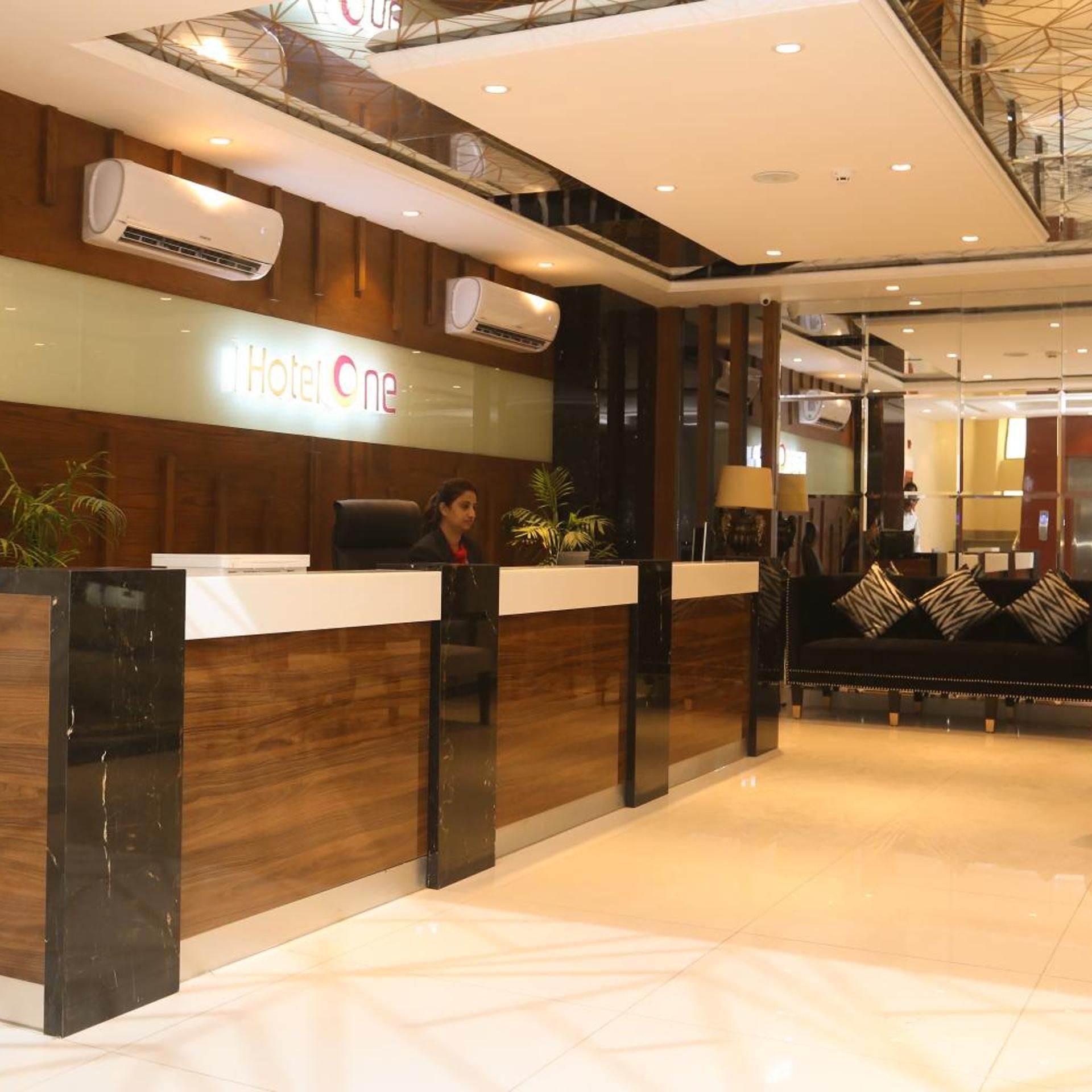 Bancada de recepção de hotel com uma placa indicando 'Hotel One', uma recepcionista e área de recepção com sofá e luminárias.