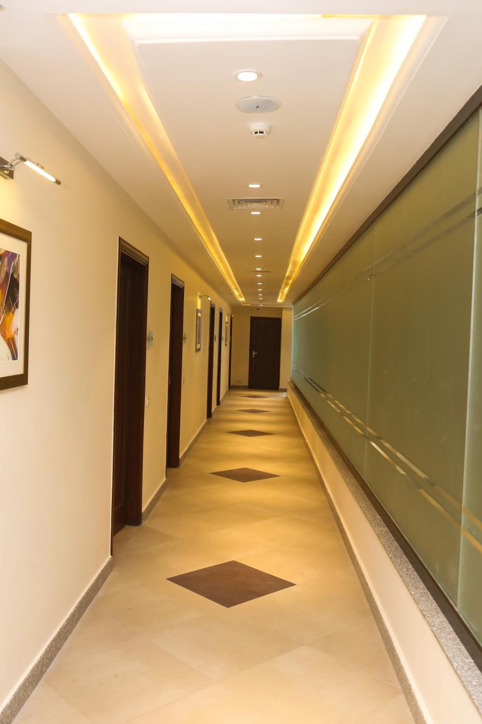 A long, narrow hallway with doors on one side and a glass wall on the other, featuring recessed lighting and patterned floor tiles.