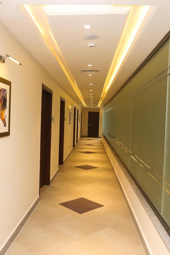 A long, narrow hallway with doors on one side and a glass wall on the other, featuring recessed lighting and patterned floor tiles.