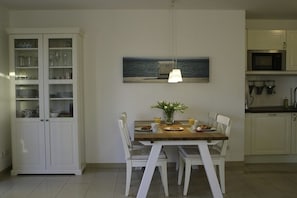 A dining area with a table set for two, a cabinet with glass doors, and a painting of a seascape. - Holiday apartment, sound of the sea (Ostseebad Nienhagen)
