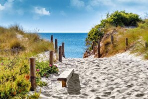 Ein Sandweg am Strand führt zum Meer, mit einer Bank und Holzpfählen.