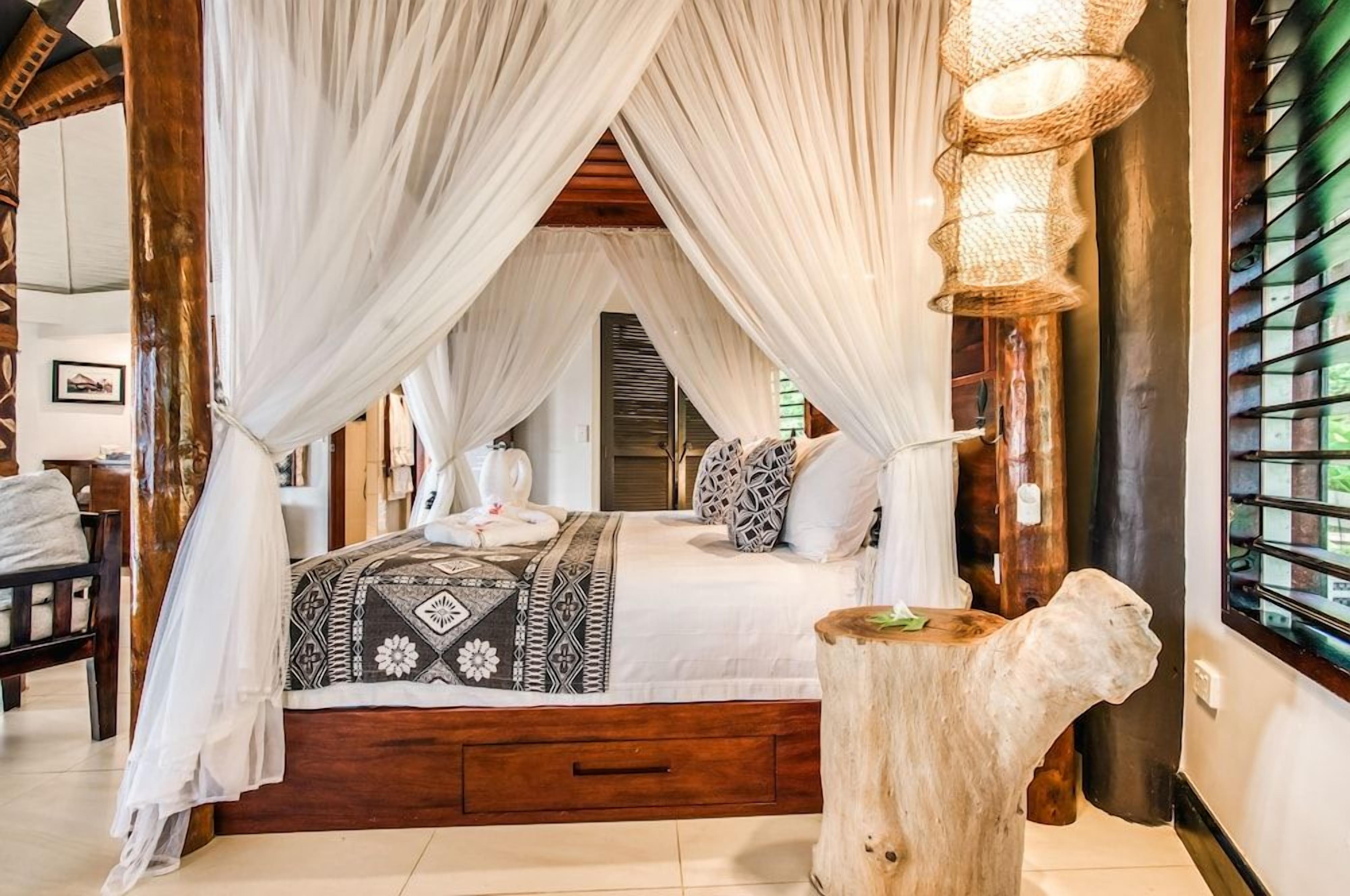 A bedroom with a canopy bed, a wooden nightstand, and a large window with blinds.