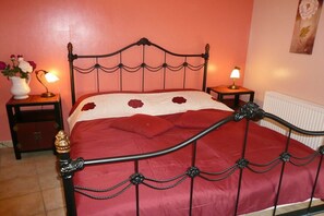 A bed with a metal frame and a red bedspread, two bedside tables with lamps, and a wall with a floral painting. - "TUSCANY" with balcony and 2 bedrooms (Waren)